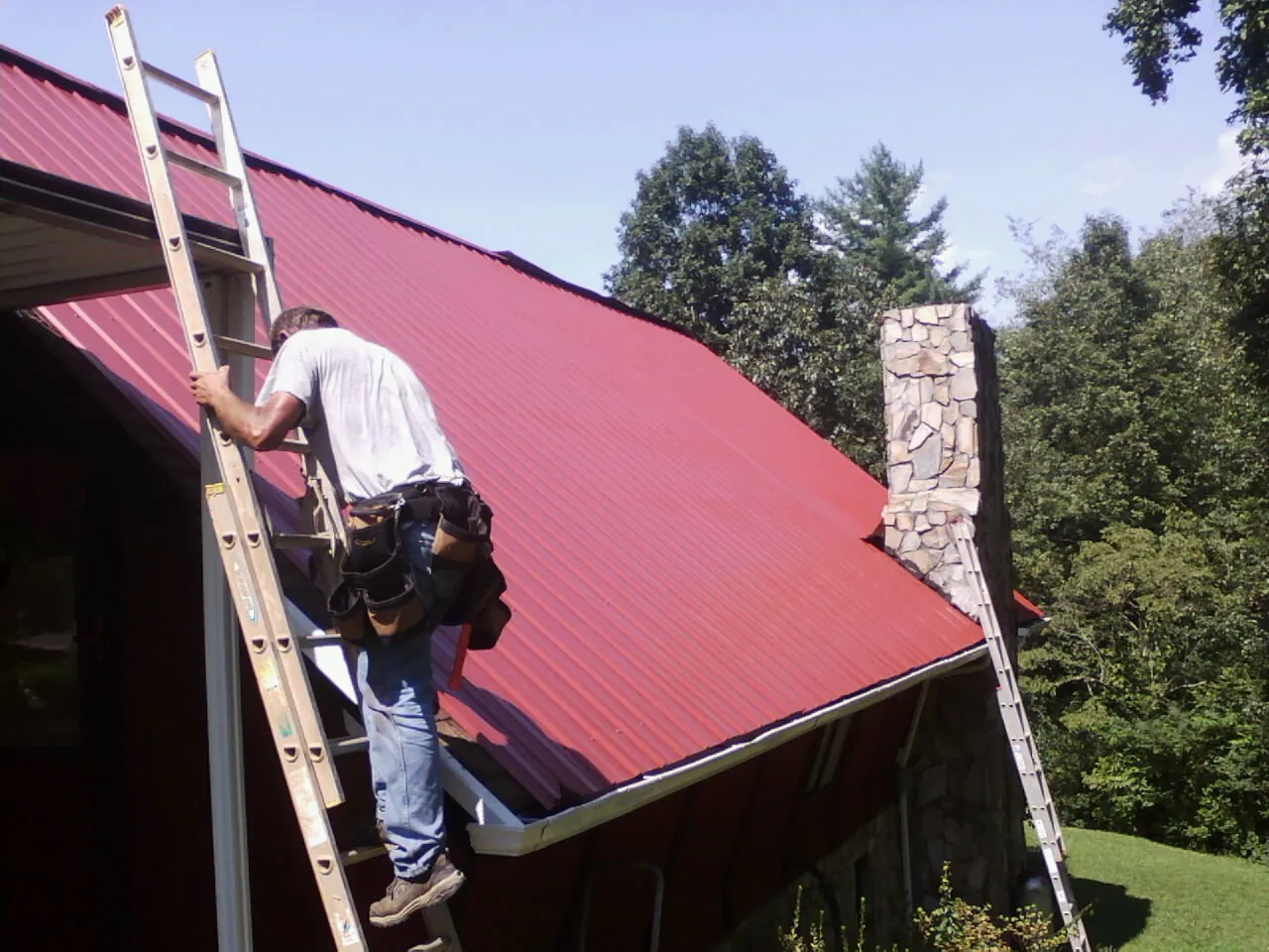 Roofer on a ladder beside a red metal roof and stone chimney — Soffit & Fascia Repair by Roofing Brothers in Salisbury Mills